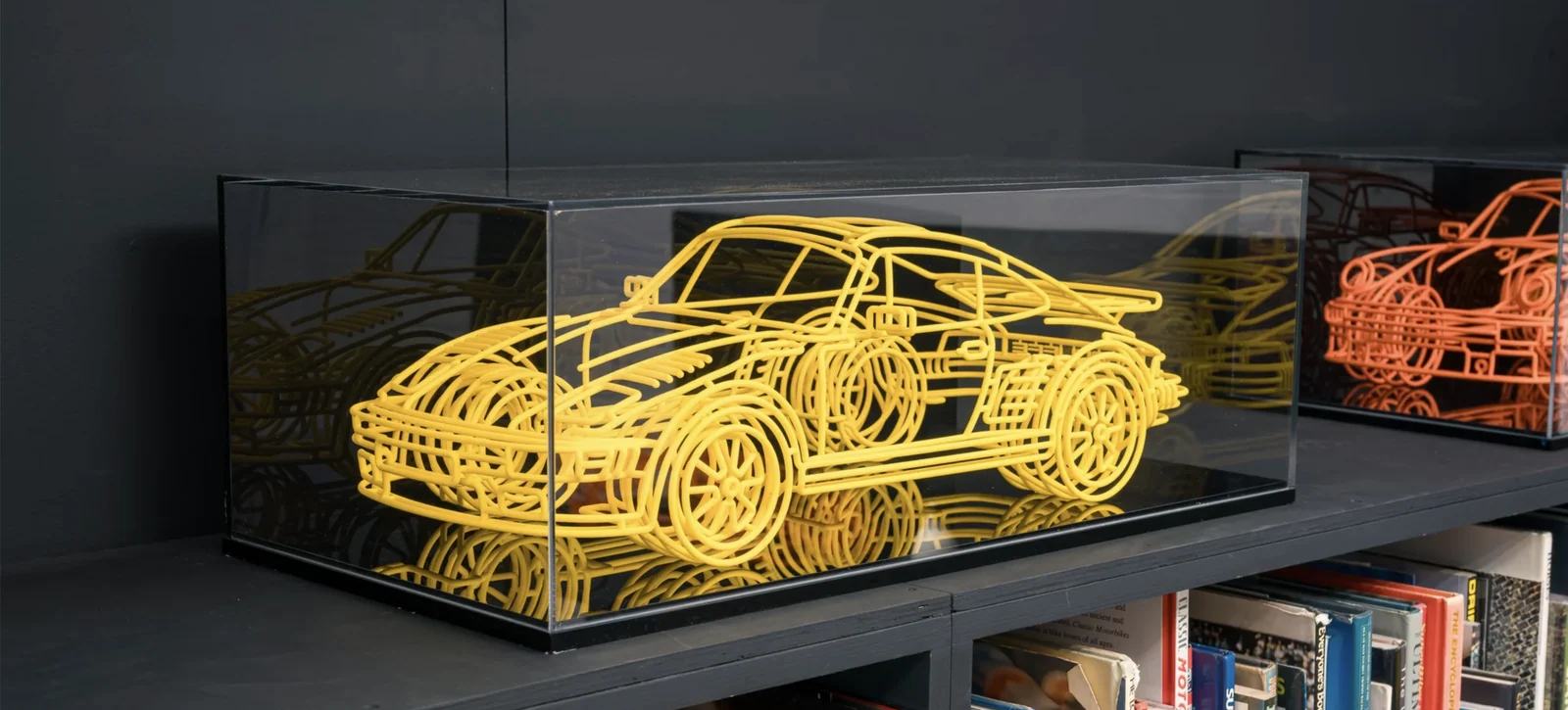 Yellow wireframe car sculpture in a glass display case, with an orange wireframe car behind it.