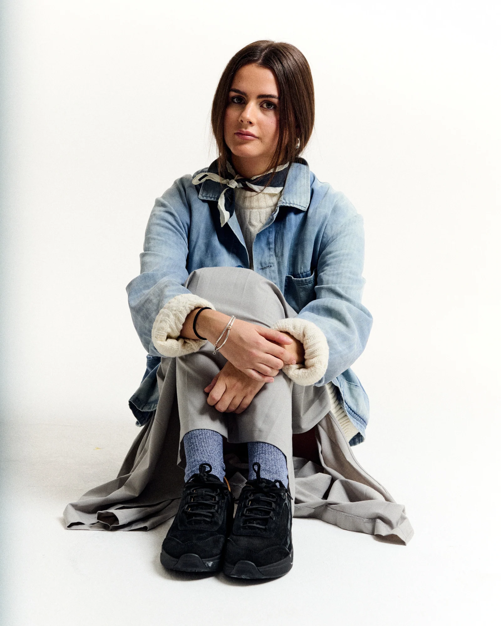 Seated model in a denim jacket with a scarf and black shoes