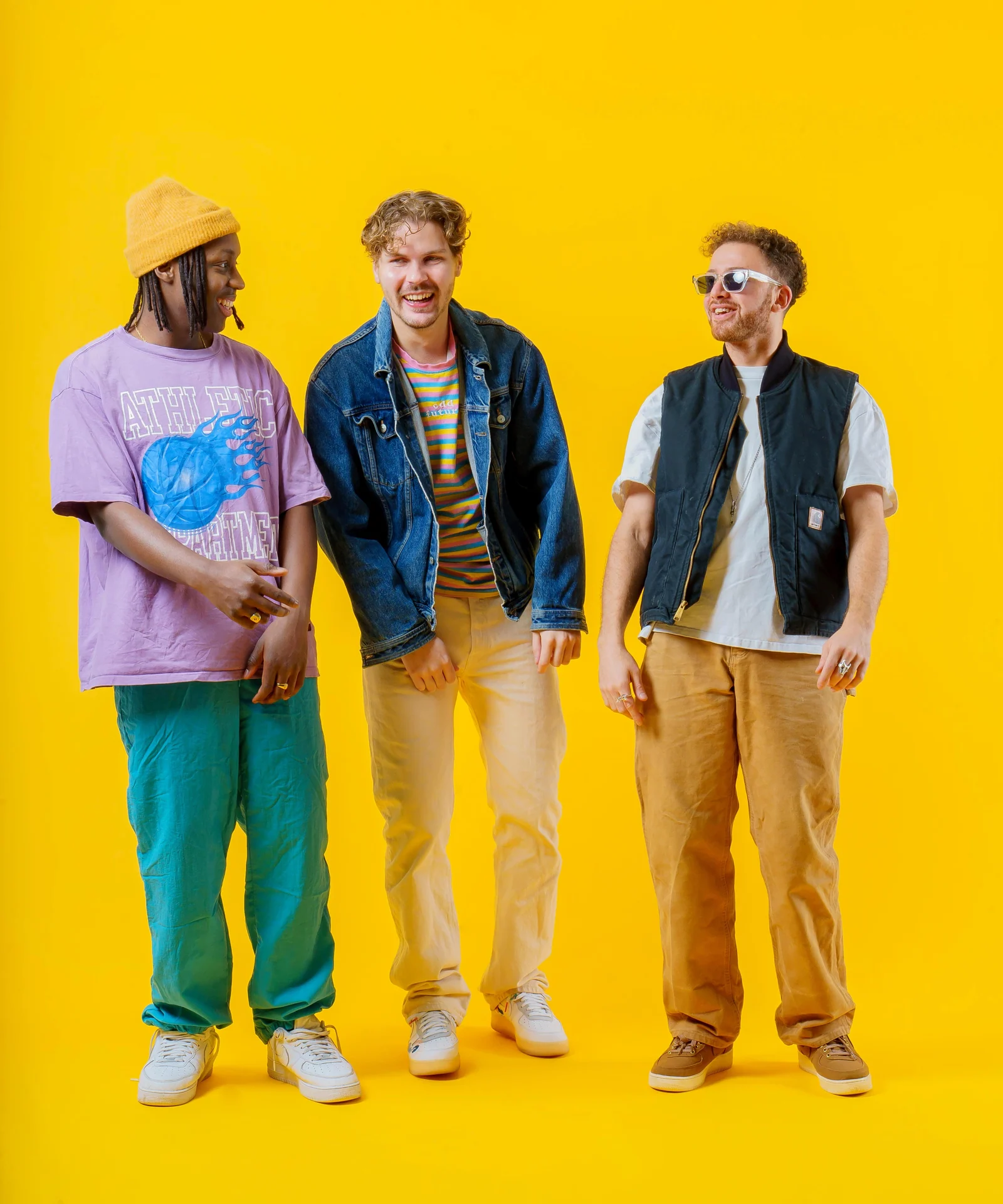 Three musicians posing in colourful outfits against a yellow studio backdrop.