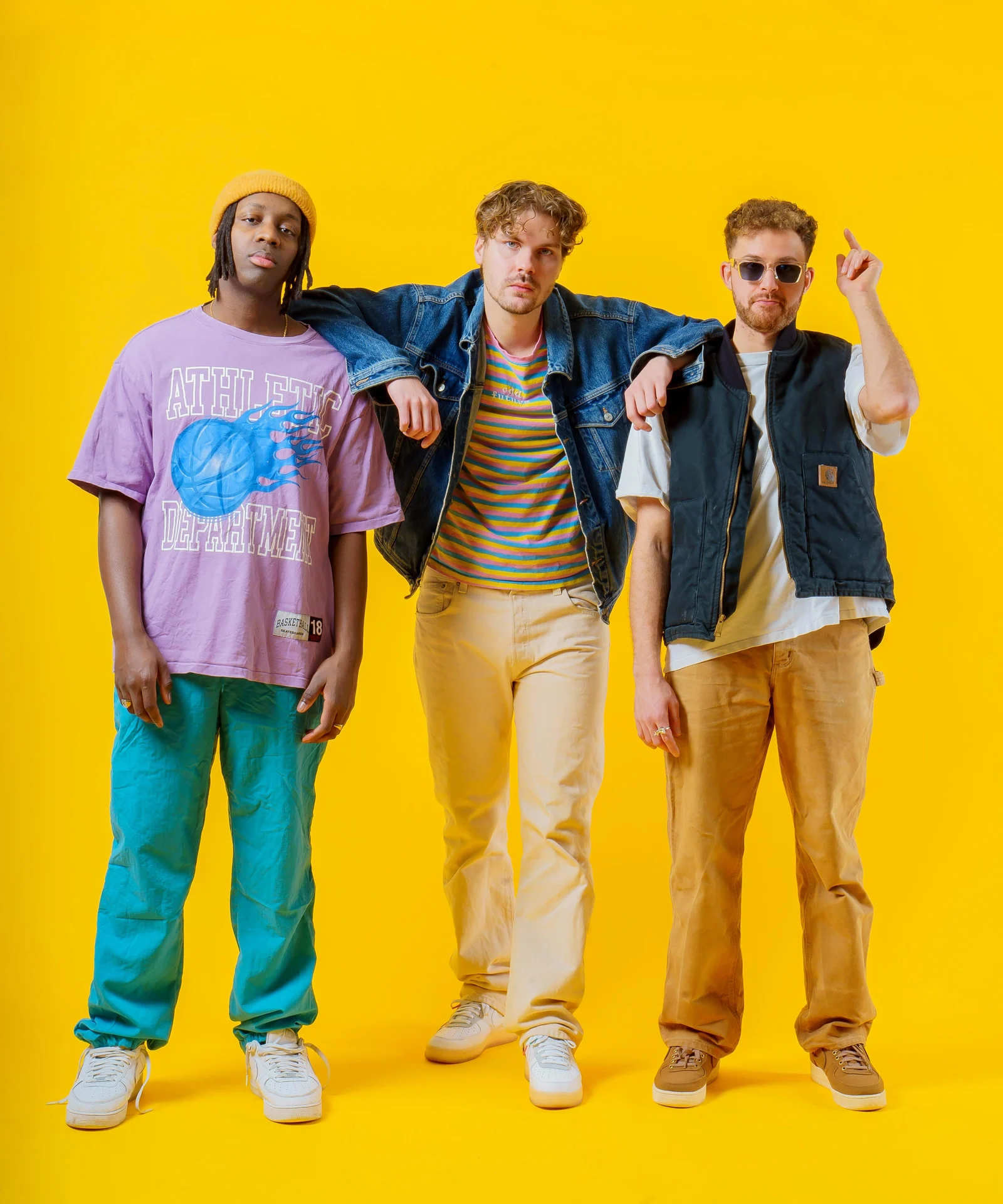 Three musicians linking arms and gesturing against a yellow backdrop.