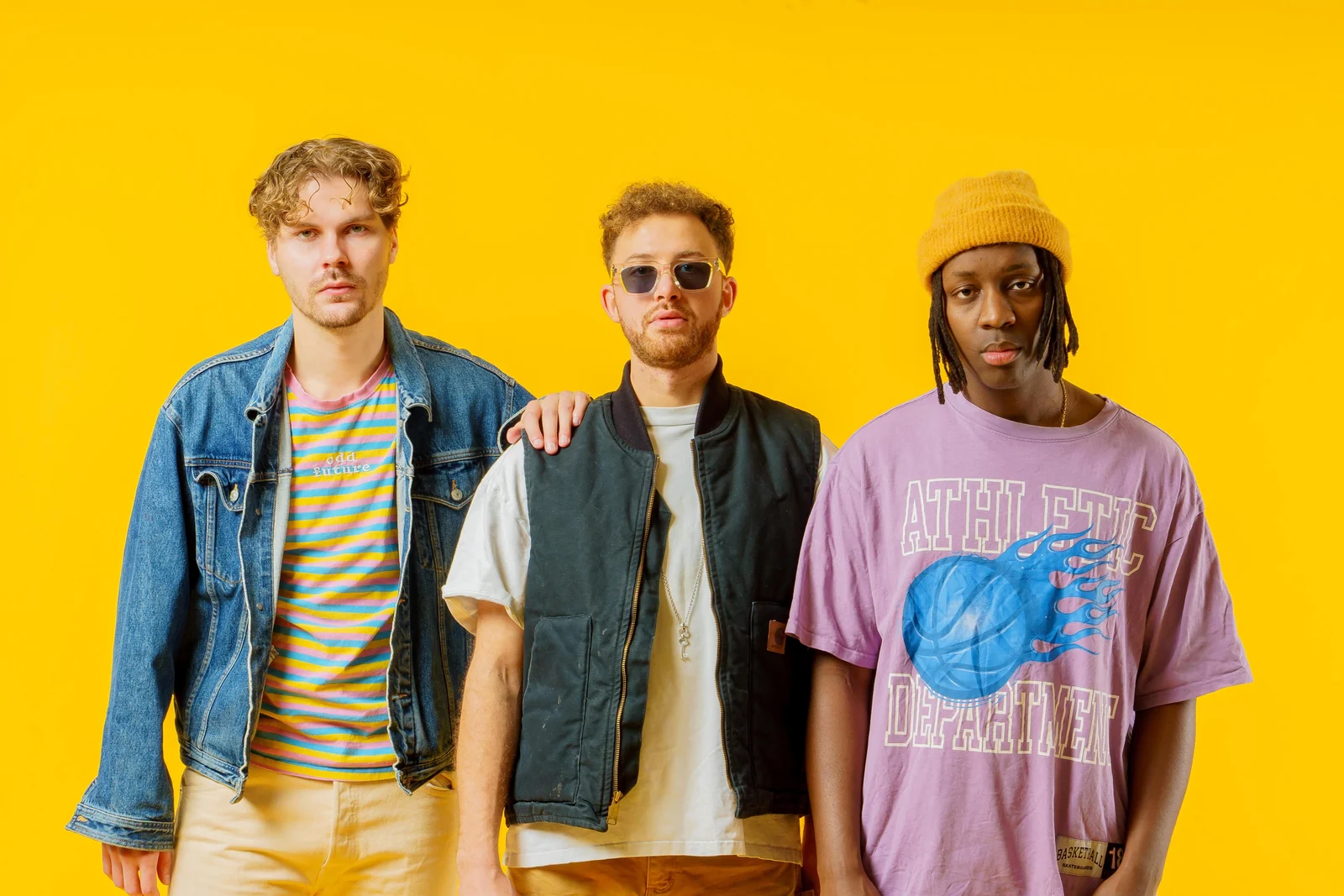 Waist-up portrait of three musicians in denim, a pale vest and a purple T-shirt against yellow.