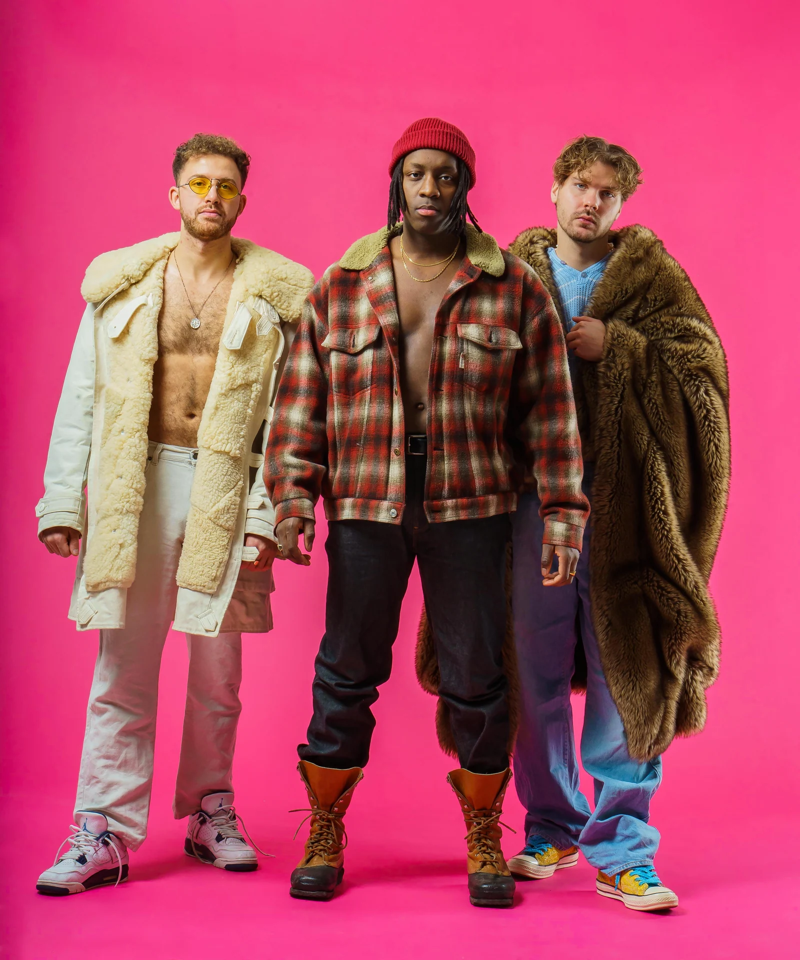 Three musicians in layered coats and a red knit cap against a pink studio backdrop.