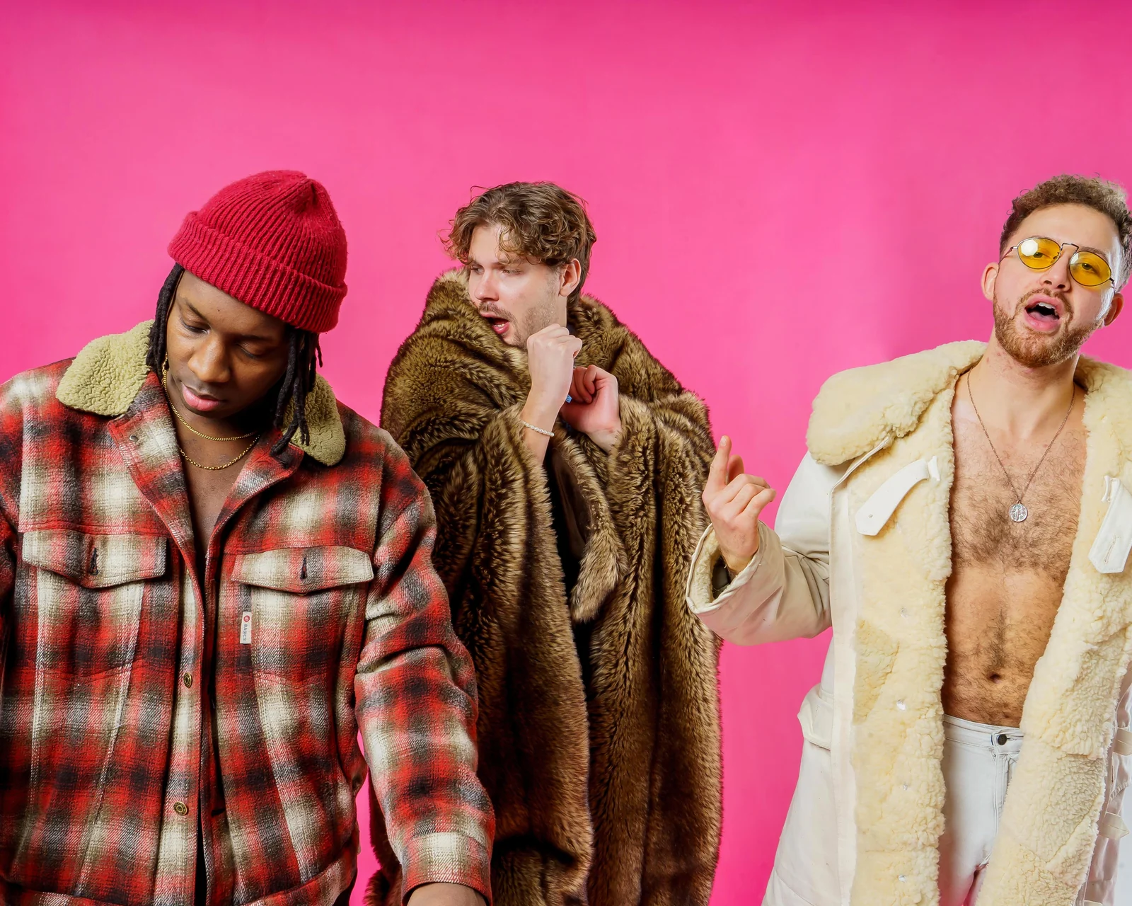 Close portrait of three musicians in coats and a red knit cap against pink.