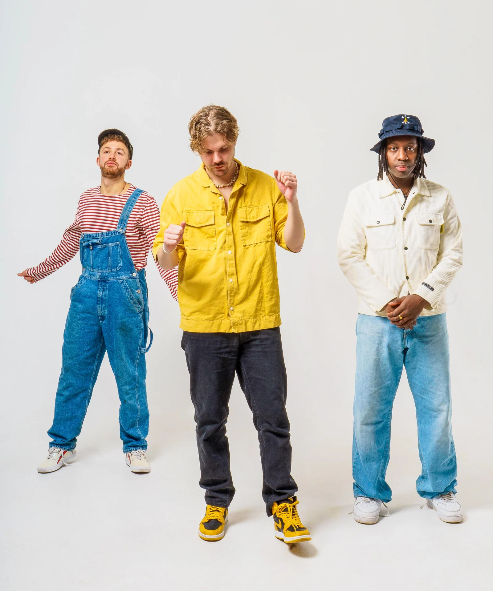 Three musicians in denim overalls, a yellow jacket and a cream shirt against white.