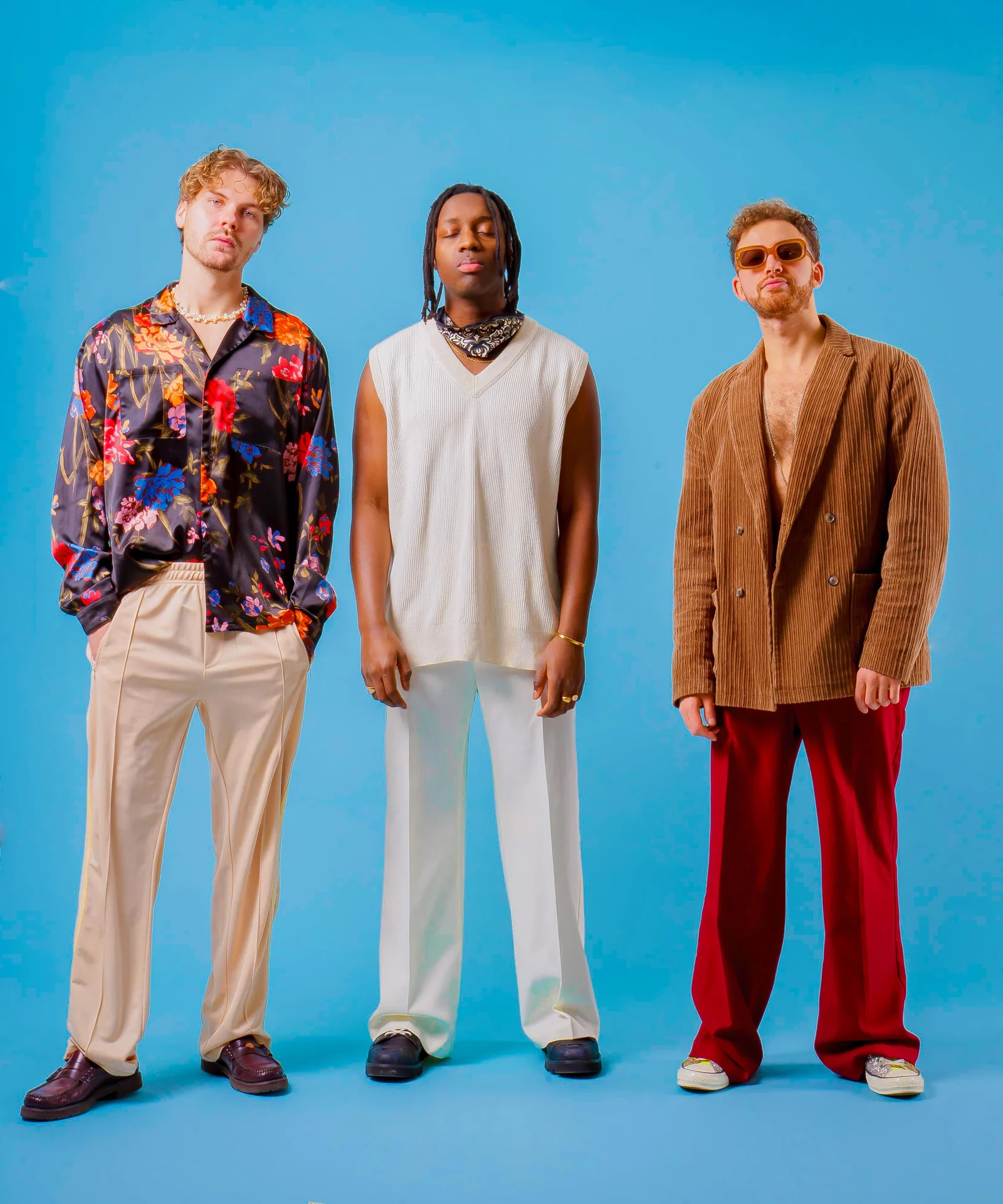 Three musicians in a floral shirt, cream knitwear and a brown blazer against blue.