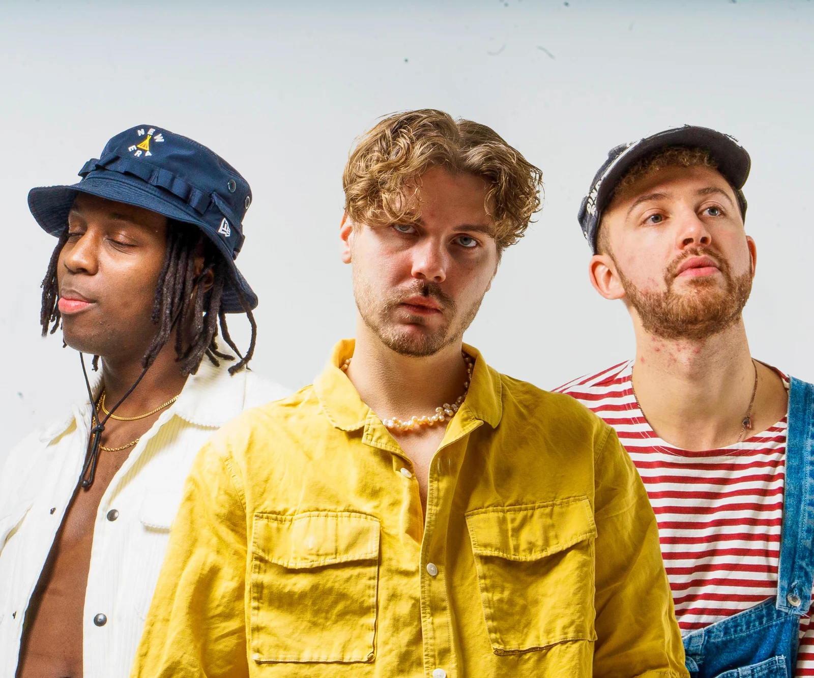 Close portrait of three musicians in bucket hats, a yellow jacket and a striped top against white.