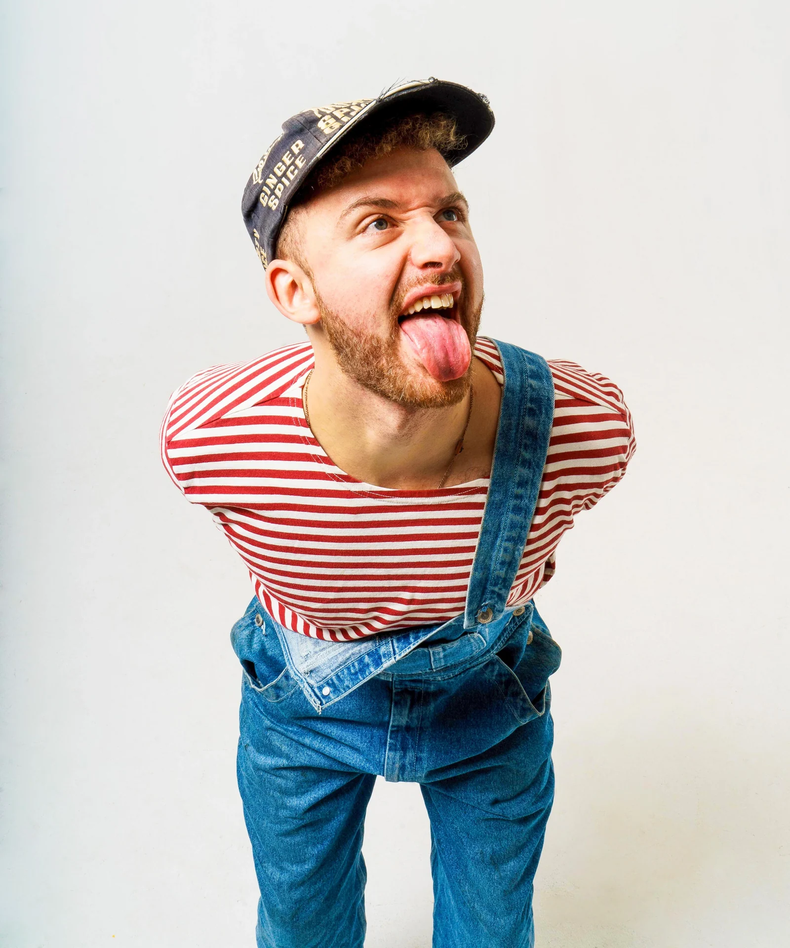 Musician in a striped top and denim overalls leaning towards the camera with his tongue out.