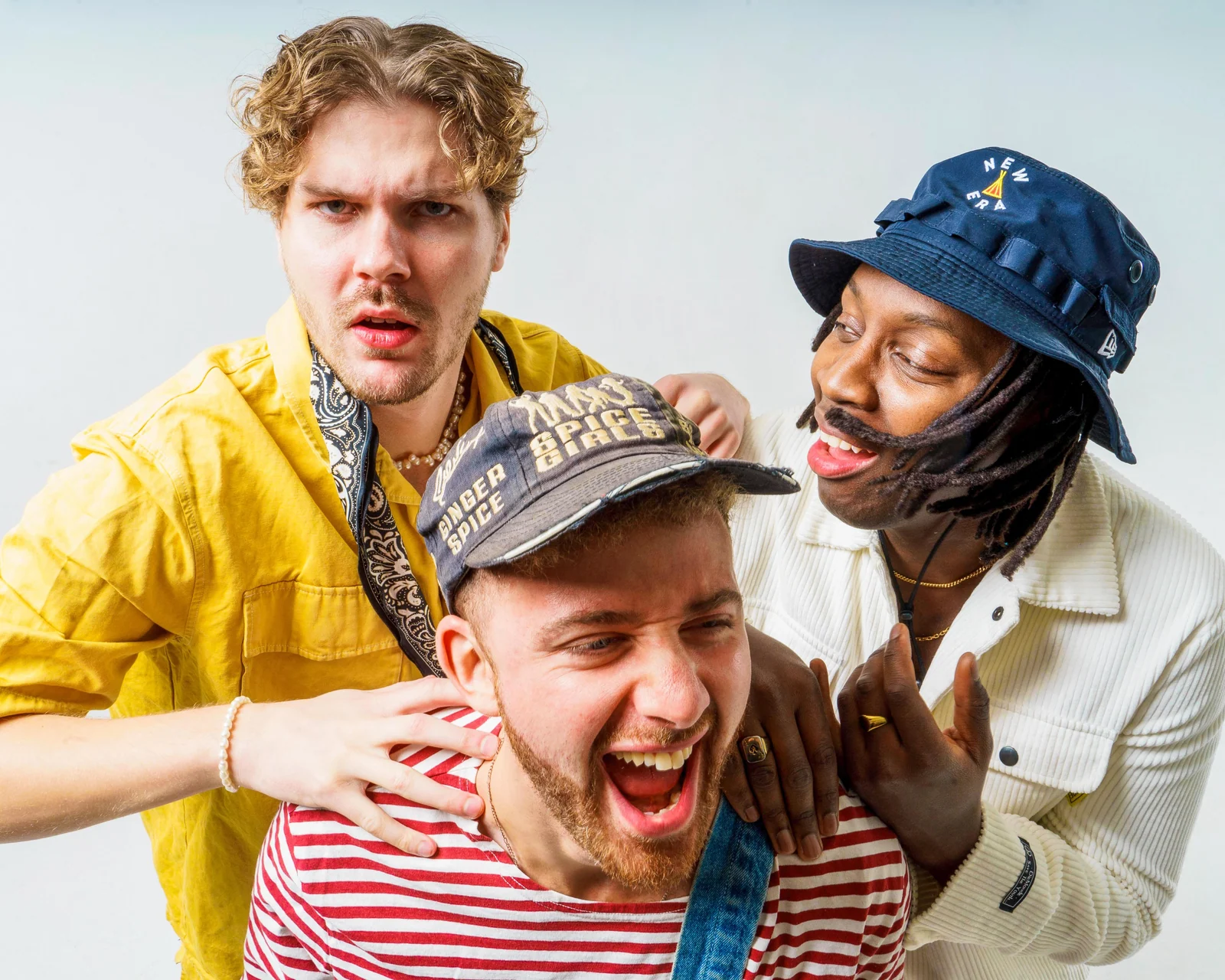 Three musicians laughing in a white studio, with one wearing a striped top and denim overalls.